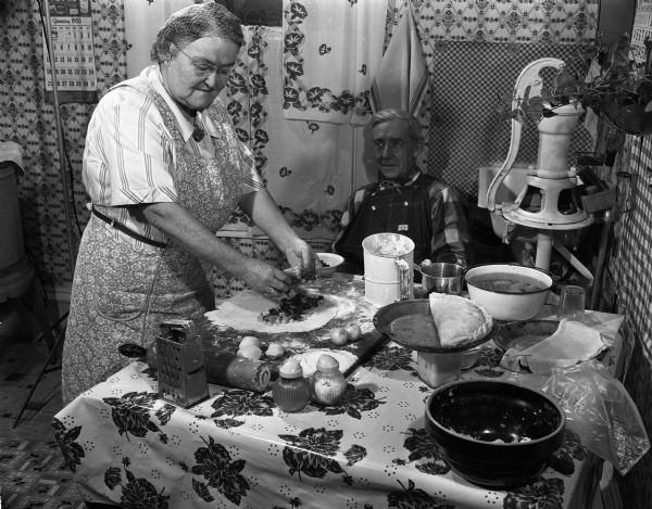 Woman wearing an apron fills a pasty in her Iowa County, Wisconsin kitchen. A man wearing coveralls is seated at the table watching. (Courtesy of <a href=" https://www.wisconsinhistory.org/Records/Image/IM58386/" target="_blank" rel="noopener noreferrer">Wisconsin Historical Society</a>)